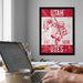 a framed poster of a red and white design with a person holding a flag, along with the text "UTAH EST. 1850 UTES". The poster is hanging on a wall, and there is a laptop and a coffee cup on a desk in the foreground.