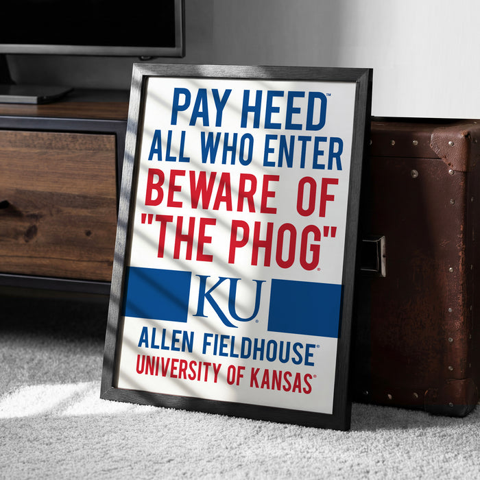 A framed sign with the text "PAY HEED ALL WHO ENTER BEWARE OF THE PHOG" and "ALLEN FIELDHOUSE UNIVERSITY OF KANSAS" on it, placed on a carpeted floor next to a wooden dresser.