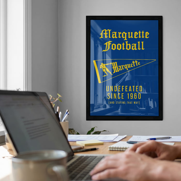 a framed poster of Marquette Football with a person working on a laptop at a desk in the foreground.