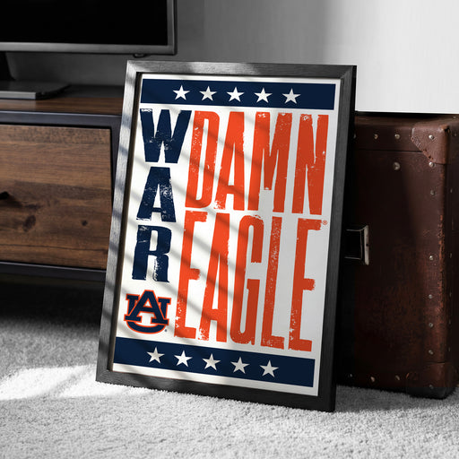 A framed poster with the text "WARM EAGLE" and the Auburn University logo, placed on a carpeted floor next to a wooden chest.