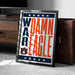 A framed poster with the text "WARM EAGLE" and the Auburn University logo, placed on a carpeted floor next to a wooden chest.