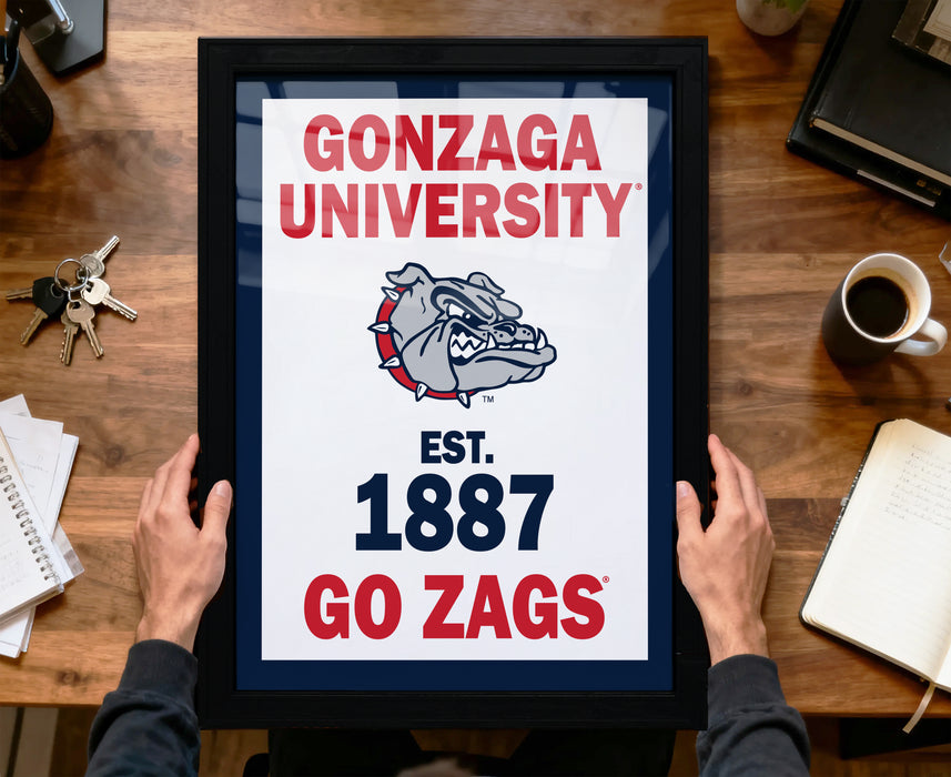 A framed poster with the text "GONZAGA UNIVERSITY EST. 1887 GO ZAGS" is being held by a person's hands, with a desk and various items in the background.