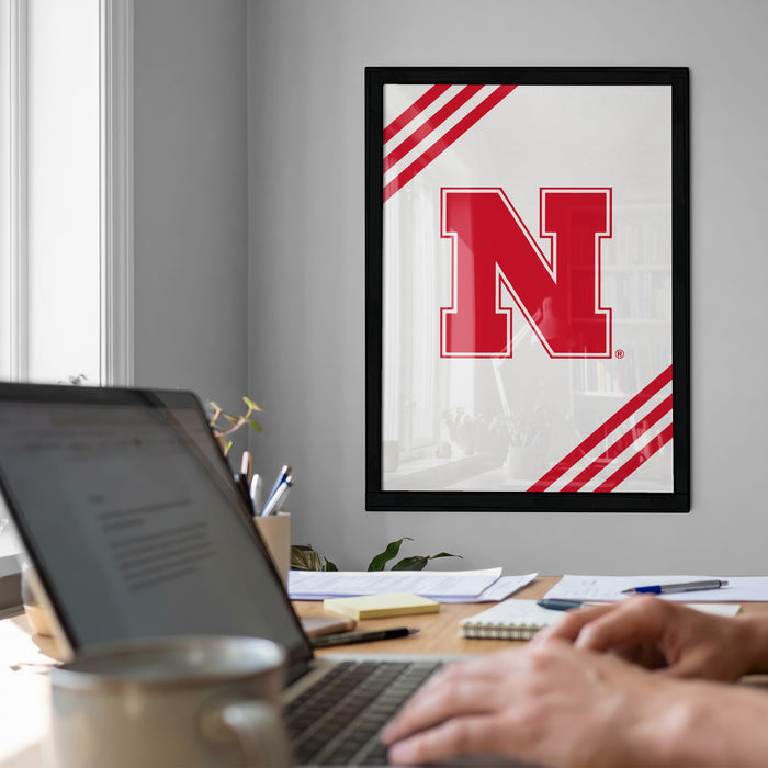 a person working on a laptop computer at a desk, with a framed poster featuring the letter 'N' hanging on the wall behind them.
