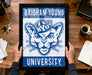 A framed poster of the University of Utah's mascot is being held by a person's hands, with a wooden desk and various items such as keys, a coffee cup, and a notebook in the background.