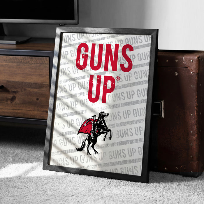A framed poster with the text "GUNS UP" prominently displayed, along with a graphic of a person riding a horse. The poster is placed on a carpeted floor next to a wooden dresser.