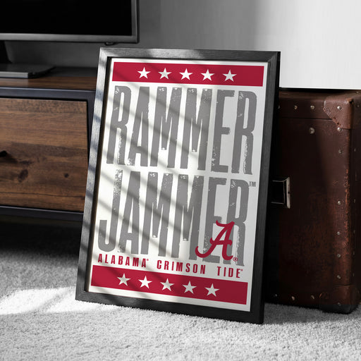 a framed poster with the text "RAMMER JAMMER" and "ALABAMA GRIMSON TIDE" printed on it, placed on a carpeted floor next to a wooden chest.