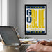 a framed poster with the text "GO BLUE MICHIGAN WOLVERS" on it, hanging on a wall above a desk with a laptop and other items on it.