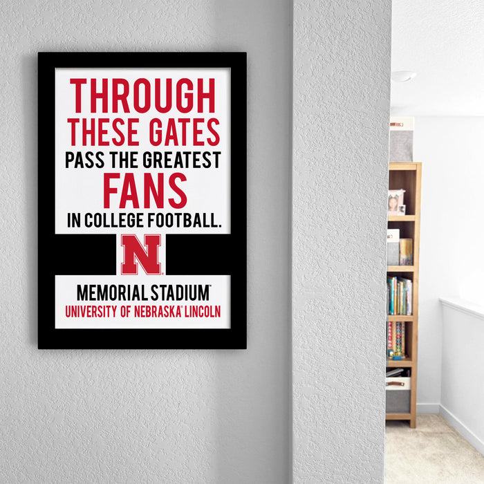 a framed sign with the text "THROUGH THESE GATES PASS THE GREATEST FANS IN COLLEGE FOOTBALL. MEMORIAL STADIUM UNIVERSITY OF NEBRASKA LINCOLN" on a wall.