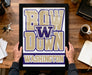 A framed poster with the text "BOW W down Washington" is being held by a person's hands, with a wooden desk and various items such as keys, a cup, and a notebook visible in the background.