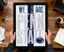 A framed poster with the text "WE ARE PENN STATE" is being held by a person's hands, with a wooden desk and various items such as keys, a cup, and a notebook in the background.