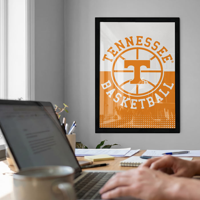 a person working on a laptop at a desk, with a framed poster of the Tennessee Basketball logo hanging on the wall behind them.