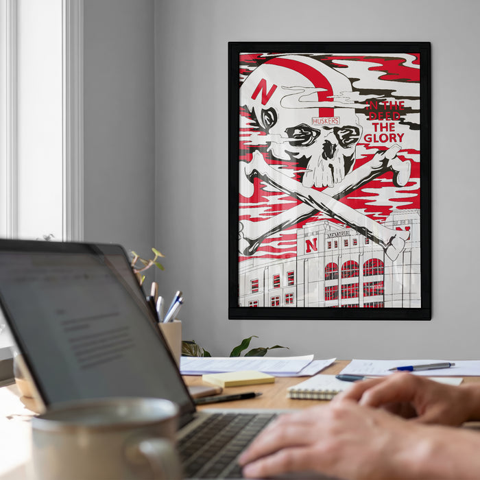 a framed poster with a skull and crossbones design, hanging on a wall above a desk with a laptop and other office supplies.