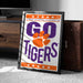 A framed poster with the text "GO TIGERS" and an orange paw print design, placed on a carpeted floor next to a wooden chest.