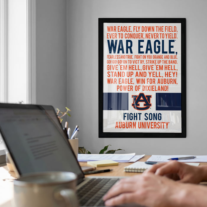 a framed poster with the text "WAR EAGLE, FEARLESS AND TRUE, FIGHT ON YOU ORANGE AND BLUE, GIVE 'EM HECKLING, GIVE 'EM HELL, STAND UP AND YELL, HELL, WIN FOR AUBURN, POWER OF DIXIELAND!" The poster is displayed on a wall above a desk with a laptop and other items.