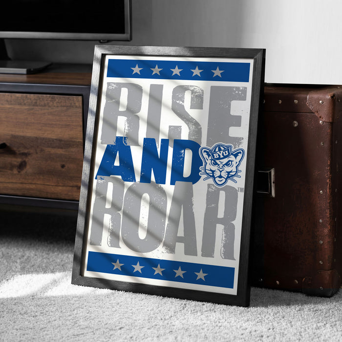 A framed poster with the text "RISE AND Roar" and a cat logo, placed on a carpeted floor next to a wooden chest.
