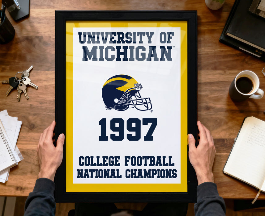 A framed poster titled "UNIVERSITY OF MICHIGAN 1997 COLLEGE FOOTBALL NATIONAL CHAMPIONS" is being held by a person's hands.