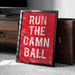 A framed poster with the text "RUN THE DAMN BALL" in white letters against a red background.