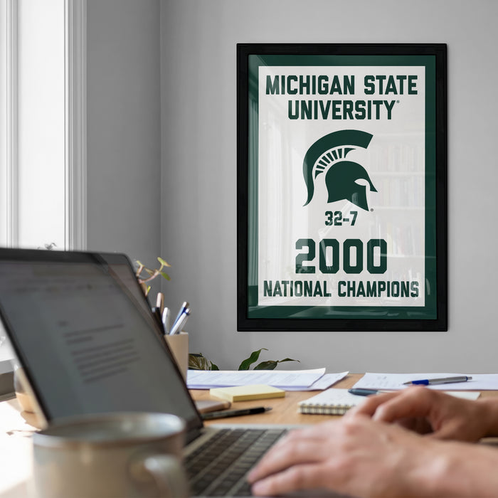 a framed poster of Michigan State University celebrating their 2000 National Champions, with a person's hands typing on a laptop in the foreground.