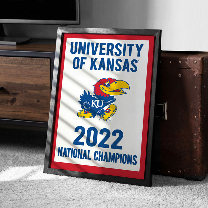 A framed poster with the text "UNIVERSITY OF KANSAS" and "2022 NATIONAL CHAMPIONS" on it, along with a graphic of a bird, is placed on the floor next to a wooden chest.