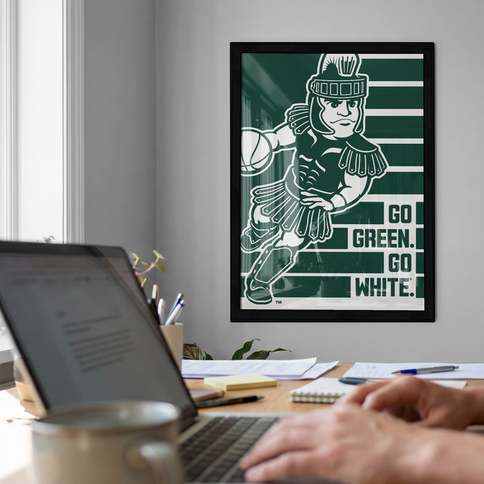a framed poster of a character holding a basketball, with the text "GO GREEN. GO WHITE." displayed below the poster. In the foreground, there is a laptop computer and a coffee mug on a desk.
