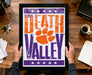 A framed poster with the text "Welcome to Death Valley" and an orange paw print graphic, placed on a wooden desk with various items such as a coffee cup, keys, and a notebook.