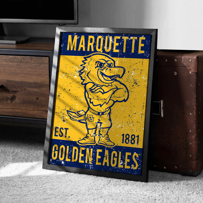 A framed poster of a cartoon eagle with the text "MARQUETTE EST. 1881 GOLDEN EAGLES" on it, placed on a carpeted floor next to a wooden chest.