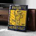 A framed poster of a cartoon eagle with the text "MARQUETTE EST. 1881 GOLDEN EAGLES" on it, placed on a carpeted floor next to a wooden chest.