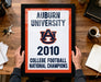 A framed sign displaying the text "Auburn University 2010 College Football National Champions" is being held by a person's hands.