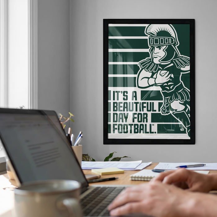 a framed poster of a football player on the wall, with the text "It's a beautiful day for football" below the poster. In the foreground, there is a laptop computer and a coffee cup on a desk, suggesting a workspace or home office setting.