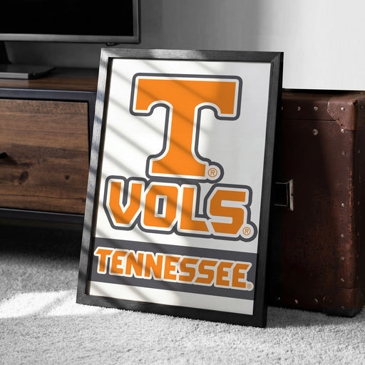 A framed poster with the word "TENNESSEE" and the logo of the University of Tennessee is displayed on a carpeted floor next to a wooden chest.