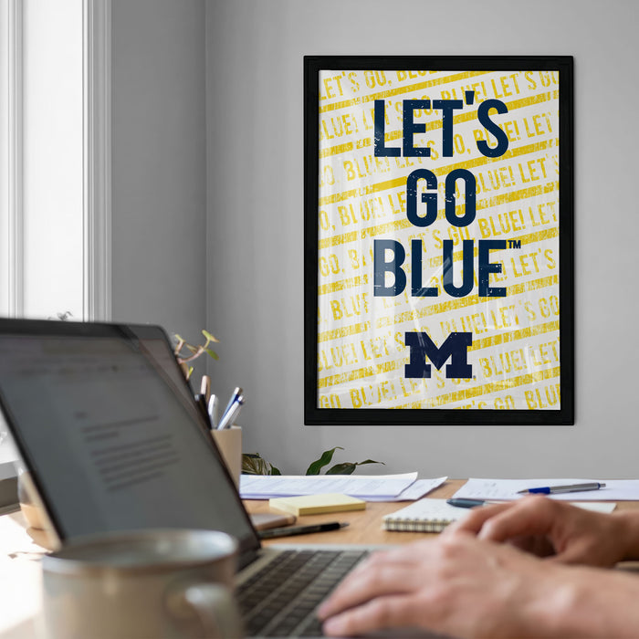 a framed poster with the text "LET'S GO BLUE" on it, which is displayed on a wall above a desk with a laptop and other items.