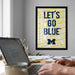 a framed poster with the text "LET'S GO BLUE" on it, which is displayed on a wall above a desk with a laptop and other items.