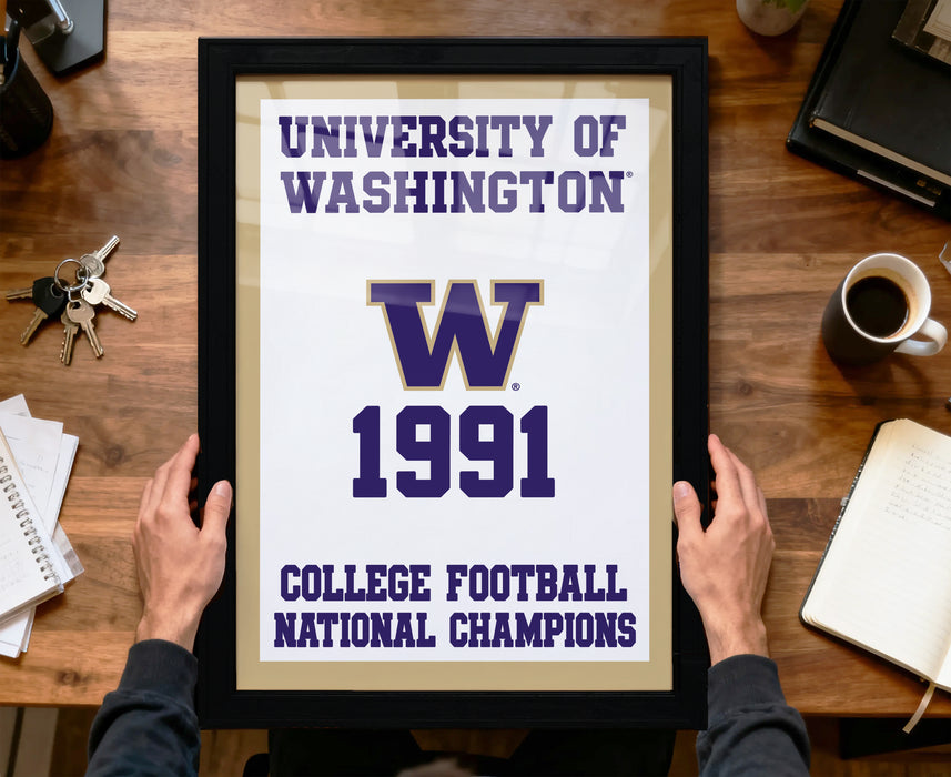 A framed poster with the text "UNIVERSITY OF WASHINGTON" and "W 1991" is being held by a person's hands, with a wooden desk and various items in the background.