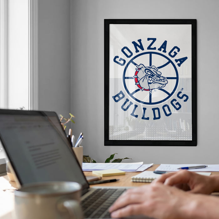 a framed poster of the Gonzaga Bulldogs on the wall, with a laptop and a coffee cup on a desk in the foreground.