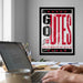 A framed poster with the text "GO TES" is displayed on the wall above a desk with a laptop and other office supplies.