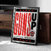 A framed poster with the text "GUNS UP" and a graphic of a gun, placed on a carpeted floor next to a wooden chest.