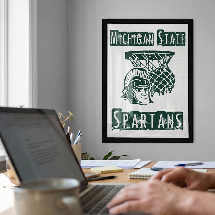 a framed poster of the Michigan State Spartans hanging on a wall, with a person's hands typing on a laptop in the foreground.