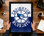 A framed poster of the Toronto Blue Jays basketball team logo is being held by a person's hands, with a desk and various items such as keys, a cup, and a notebook visible in the background.