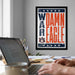 a framed poster with the text "DAMN EAGLE" on it, which is displayed on a wall above a desk with a laptop and other items.