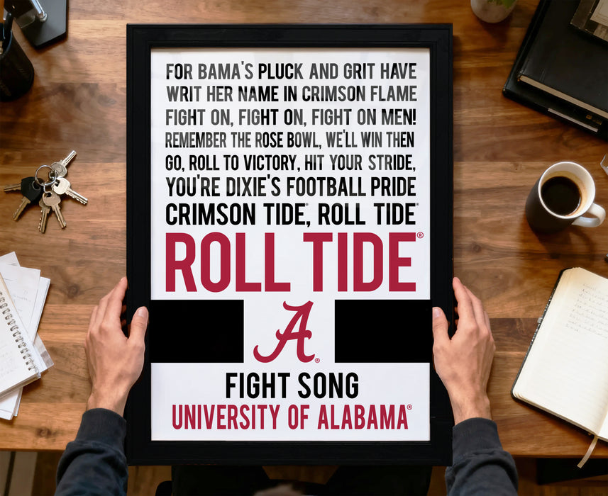 a framed poster with text that reads "For Bama's Pluck and Grit Have WRIT HER NAME IN CRIMSON FLAME FIGHT ON, FIGHT ON, FIGHT ON MEN! REMEMBER THE ROSE BOWL, WE'LL WIN THEN GO, ROLL TO VICTORY, HIT YOUR STRIDE, YOU'RE DIXIE'S FOOTBALL PRIDE CRIMSON TIDE, ROLL TIDE". The poster is being held by a person's hands, and there is a notebook, a cup, and a keychain on the desk in the background.