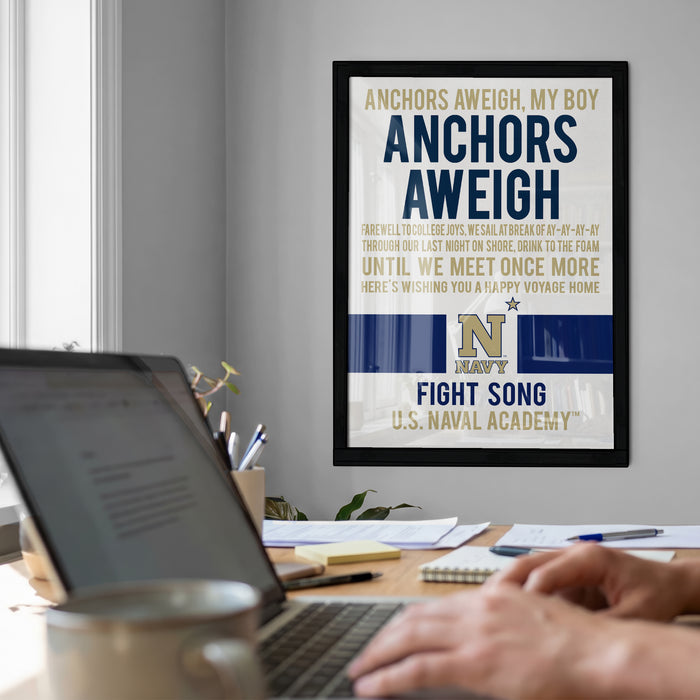 a framed poster with a humorous message about anchors and a naval school, hanging on a wall above a desk with a laptop and other items.