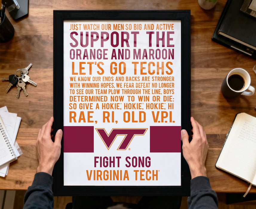 a person holding up a framed poster with text, including a Virginia Tech logo, on a wooden desk with various items such as a coffee cup, keys, and a notebook.