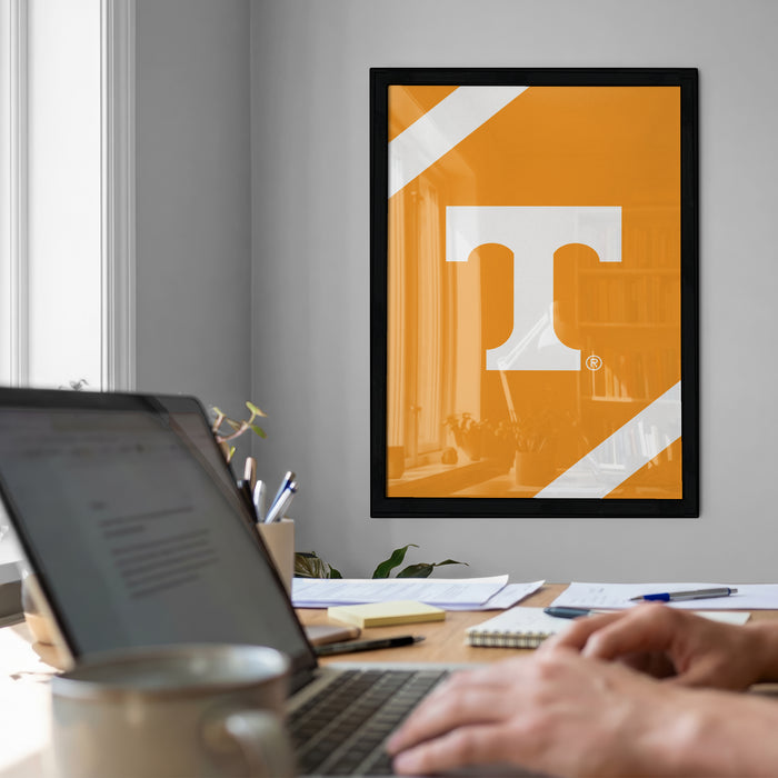 a workspace with a laptop computer, a framed poster with a large letter 'T' on the wall, and various office supplies on a desk.