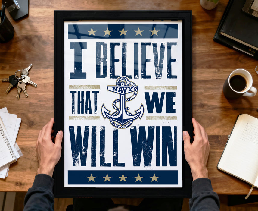 A framed poster with the text "I BELIEVE THAT WE WILL WIN" is being held by a person's hands, with a wooden desk and various items such as a cup, keys, and a notebook in the background.