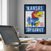 a framed poster of a Kansas Jay Hawks logo hanging on a wall, with a person's hands visible in the foreground using a laptop computer.
