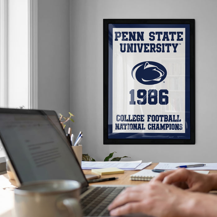 A framed poster of Penn State University from 1986 hangs on a wall, with a laptop and a coffee mug on a desk in the foreground.