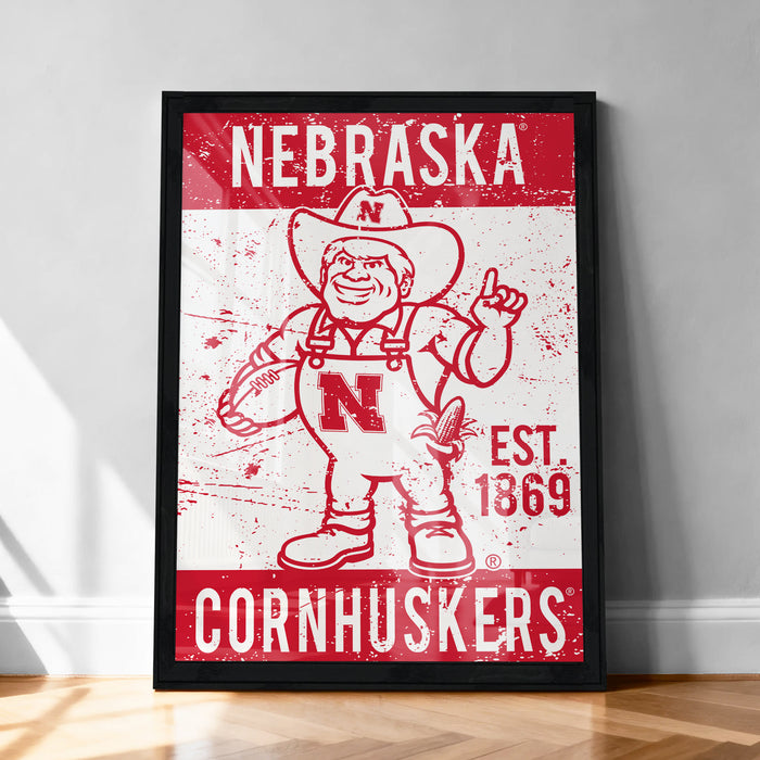A framed poster featuring the Nebraska Cornhuskers, a sports team, with a cartoon illustration of a man wearing a Nebraska hat and holding a football.