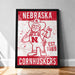 A framed poster featuring the Nebraska Cornhuskers, a sports team, with a cartoon illustration of a man wearing a Nebraska hat and holding a football.
