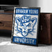 A framed poster with the text "BRIGHAM YOUNG" and "YU" is displayed on a carpeted floor next to a brown suitcase.