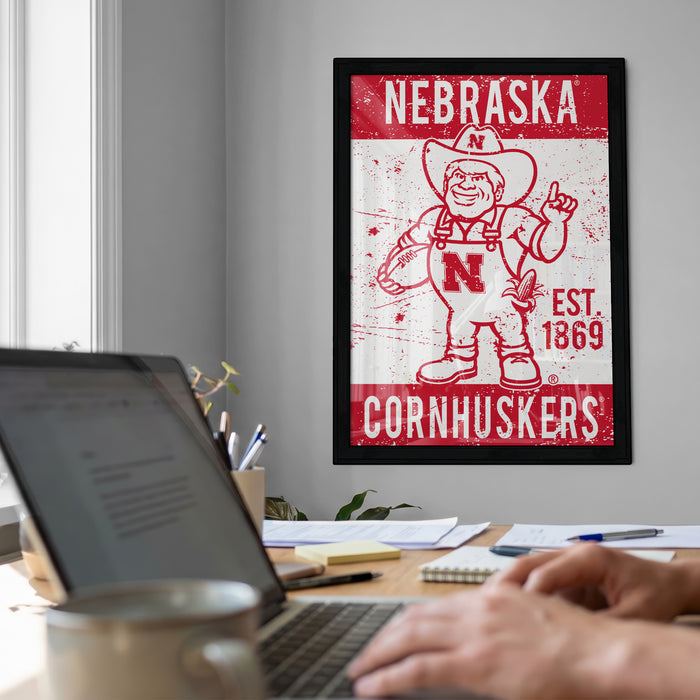 a framed poster of a cartoon character from Nebraska, with the text "NEBRASKA" and "CORNHUSKERS" prominently displayed. In the foreground, there is a laptop computer and a coffee mug on a desk, suggesting a workspace or home office setting.